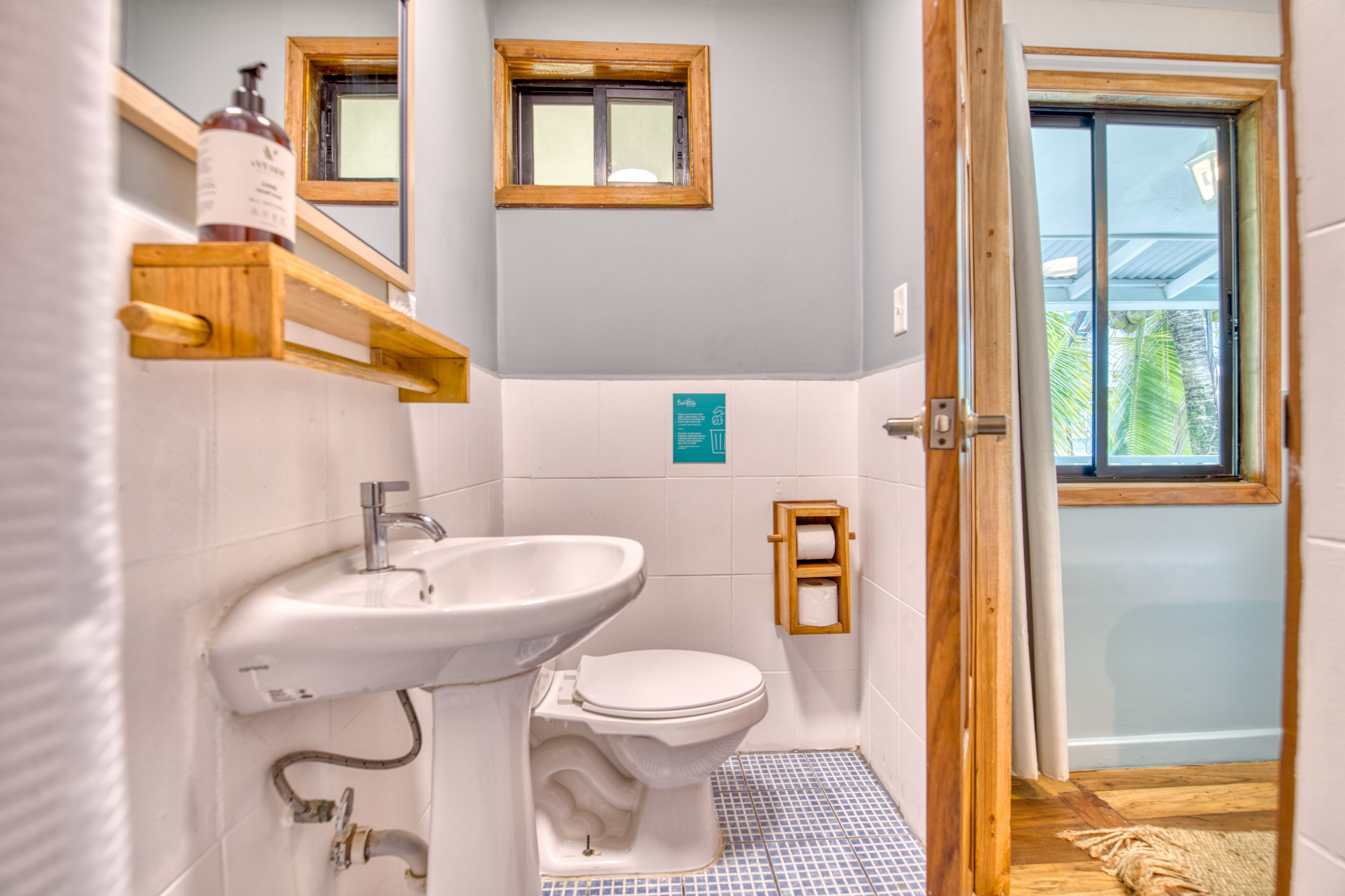 Een badkamer met een wit wastafelmeubel, toilet en een houten plank met toiletartikelen. Er is een raam boven het wastafelmeubel en een deur die leidt naar een balkon.