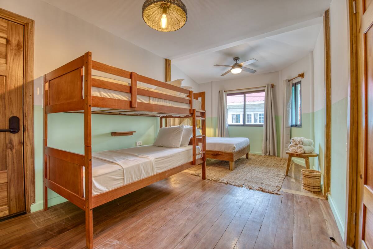 A room with a bunk bed, a single bed, a ceiling fan, and a window with curtains.