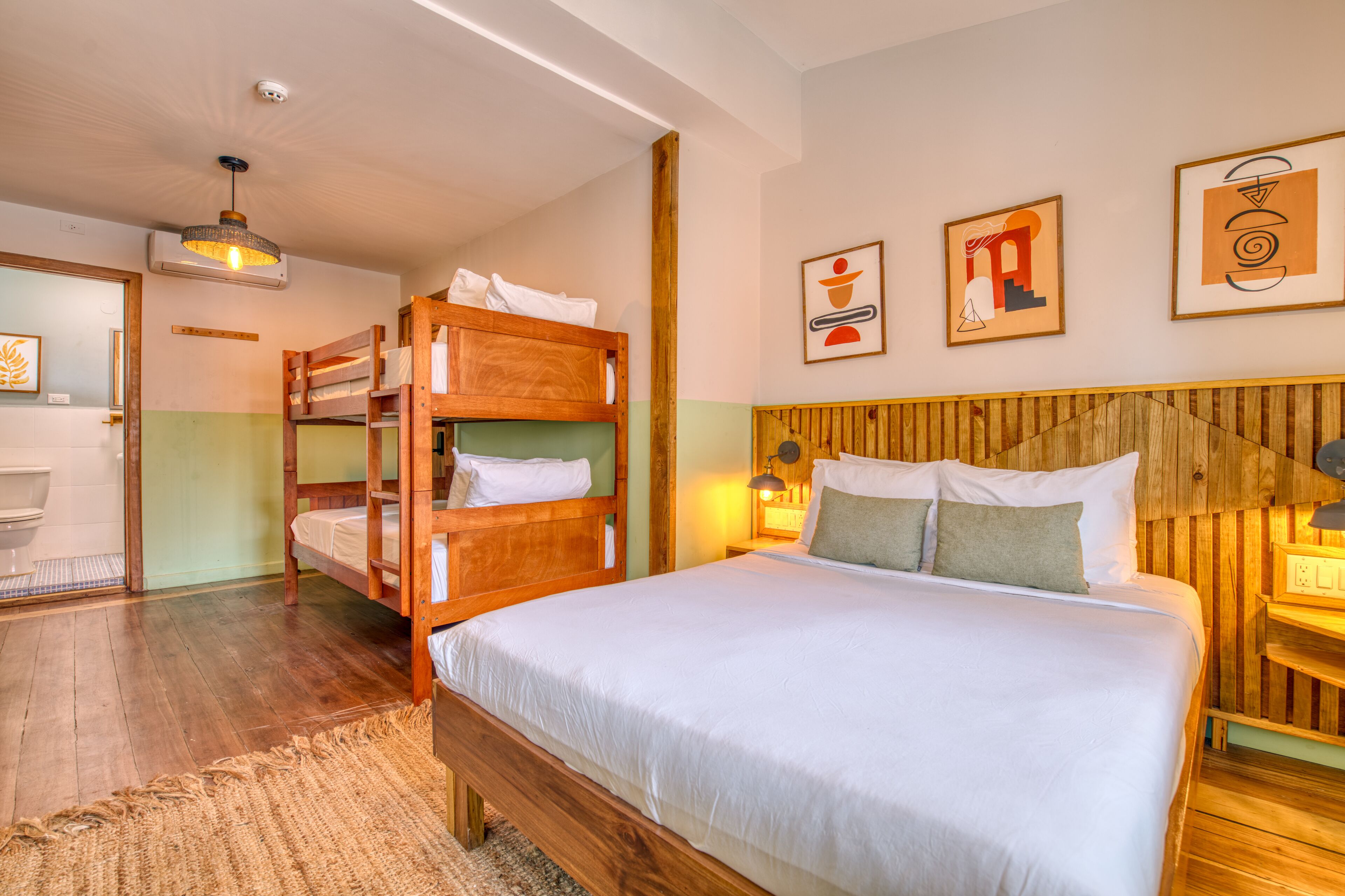 A room with a large bed, wooden bunk beds, a bathroom with a toilet, and a wooden wall with abstract art.