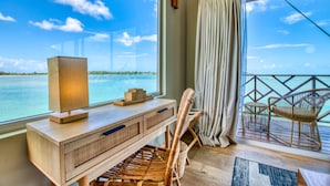 Una habitación con un ventanal grande con vistas al agua, un escritorio de madera con una lámpara, sillas de mimbre y vista al mar.