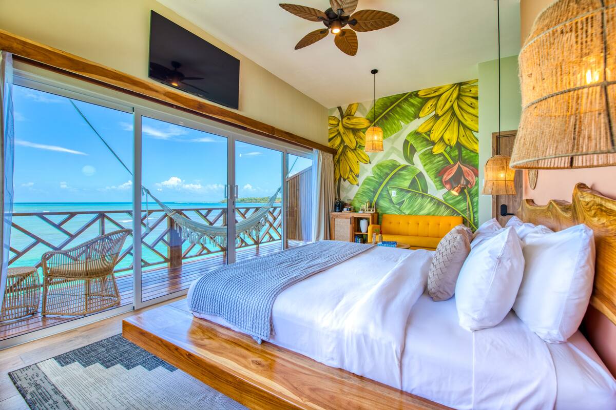 A bedroom with a large bed, a view of the ocean, a ceiling fan, and a tropical-themed wall mural.
