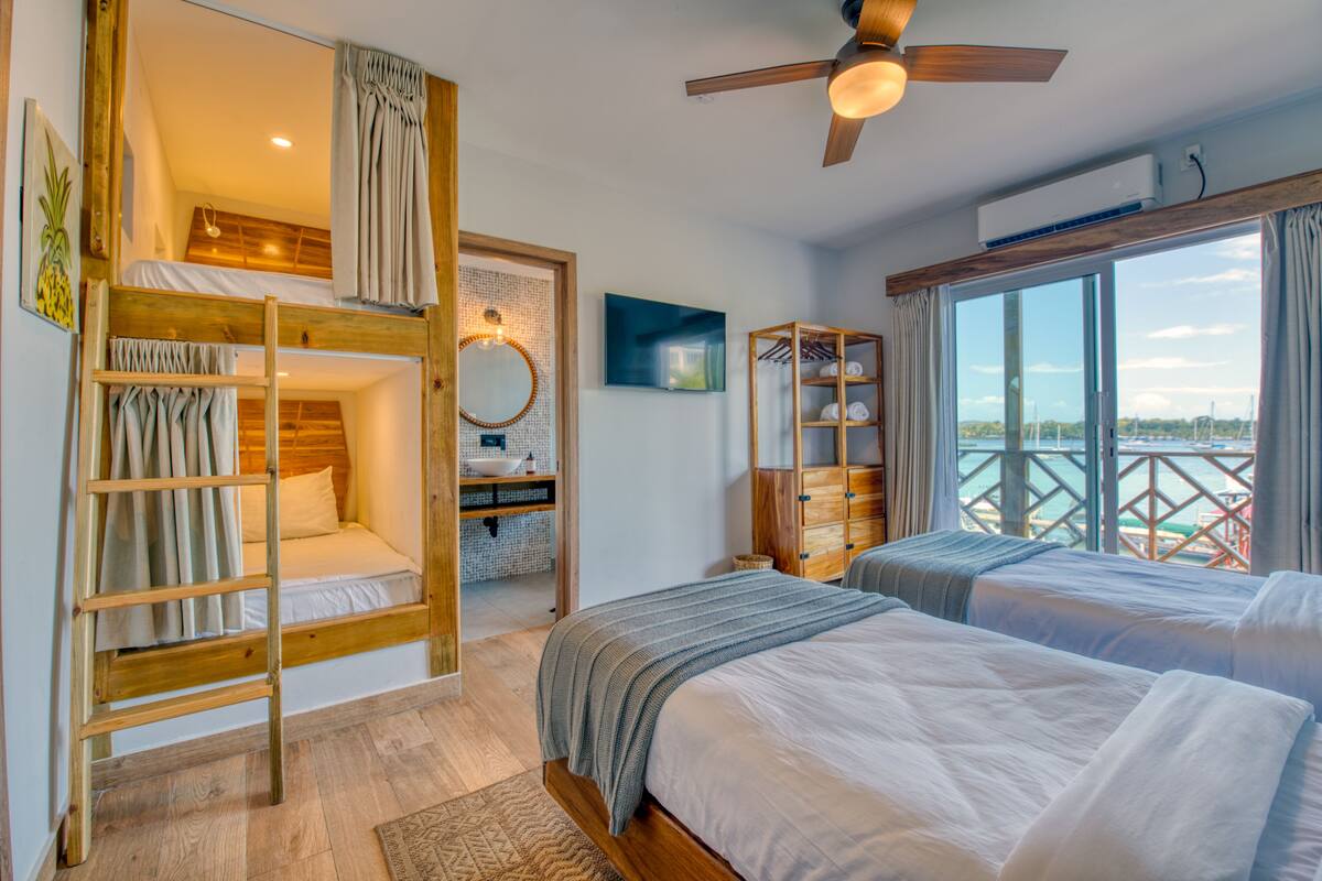 A room with two beds, a wooden ladder, a ceiling fan, a large window with a view, and a wooden wardrobe.
