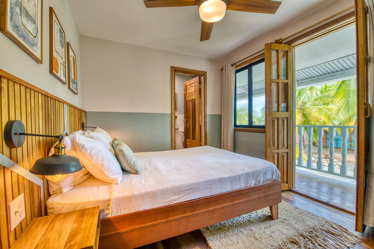 A bedroom with a wooden bed, a nightstand, a ceiling fan, and a large window with a view of palm trees.