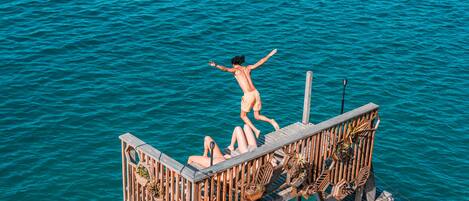 A wooden deck extending over turquoise water, with two people sitting and one person jumping.