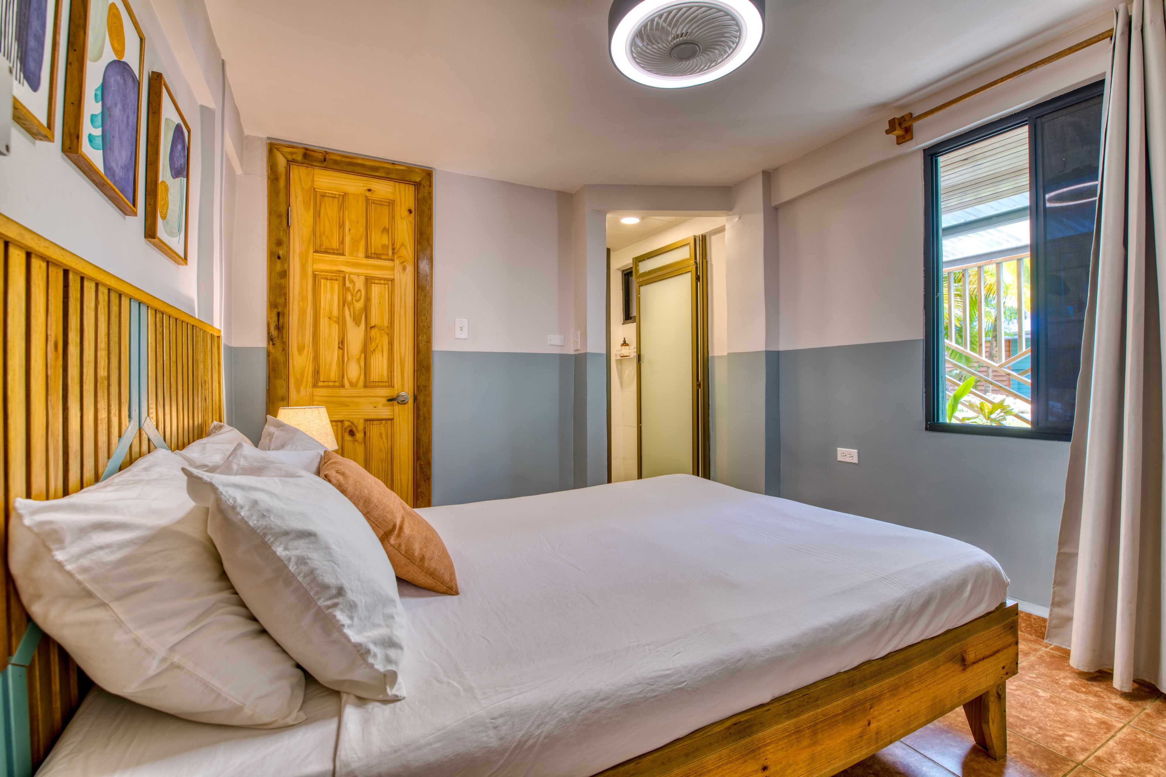 Un dormitorio con cama de madera, ropa de cama blanca, cabecero de madera, ventilador de techo, ventana con cortinas y puerta que da a otra habitación.
