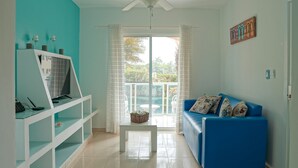 A living room with a blue sofa, a white TV stand, a ceiling fan, and a view through a glass door to the outdoors. - Ria Aparta Hotel (Puerto Plata)