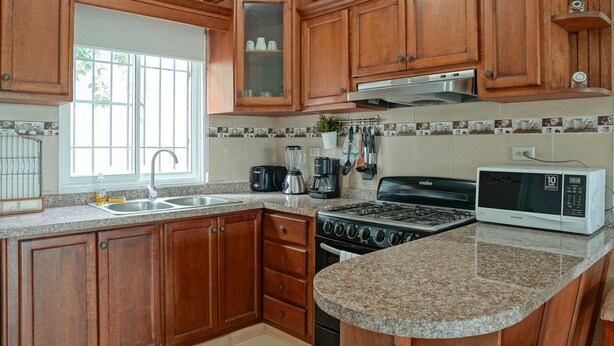 A kitchen with wooden cabinets, a gas stove, a microwave, and a window with a view of trees. - Ria Aparta Hotel (Puerto Plata)