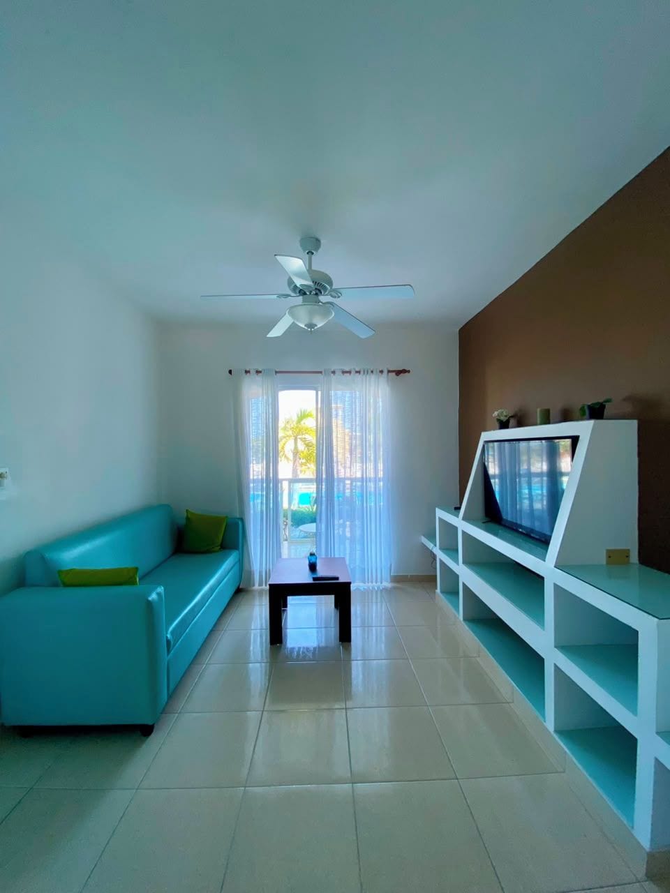 Un salon avec un canapé de couleur turquoise, un téléviseur posé sur un meuble blanc, un ventilateur de plafond et une vue sur l’extérieur, visible à travers une porte vitrée.