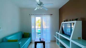 Un salón con un sofá color turquesa, un televisor sobre un soporte blanco, un ventilador de techo y vistas a través de un vidrio.