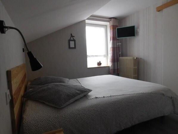 Un dormitorio con cama, escritorio, silla, ventana, radiador y televisor montado en la pared.