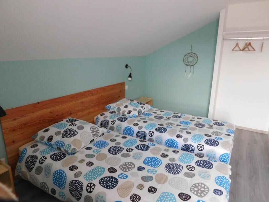 A bedroom with a wooden headboard, a bed with patterned bedding, a nightstand, a wall-mounted lamp, and a dreamcatcher.