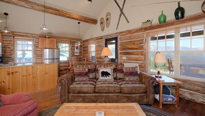 Ein Blockhauss Innenraum mit gemütlichem Wohnbereich, Holzwänden, einer Couch mit gemusterten Kissen, einem Esstisch und einer Küche mit Holzausstattung.
