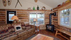 Ein Holzblockhaus-Interieur mit einer Couch, einem Tisch, einer Lampe, einem Fenster und einem Kaminofen.