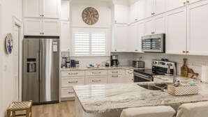 A modern kitchen with white cabinetry, a stainless steel refrigerator, a microwave, and a large island with a sink. - SEA SHANTY- Beachfront, Topnotch Comfort & Fully-Stocked! (Mexico Beach)