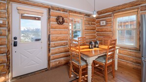A log cabin interior with a dining table, chairs, a refrigerator, and a door leading to a scenic view.