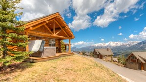 A log cabin with a wooden deck and a hot tub, surrounded by mountains and a clear sky.