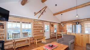 A log cabin interior with a wooden bench, a table with a chessboard, a sofa, and a kitchen area with a clock.