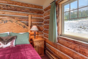 A log cabin room with a bed, a nightstand, a lamp, and a window with a snowy view. - Ski-in/Ski-out home w/private hot tub in Moonlight Basin - Cowboy Heaven Cabin - Wild West (Big Sky)