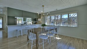 Una cocina amplia con una mesa de comedor de madera y sillas blancas, ventanas grandes con vistas a árboles, y un techo blanco con vigas de madera.