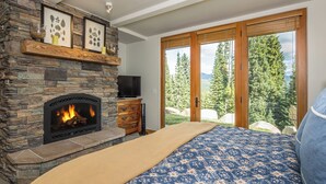 Ein Schlafzimmer mit Kamin aus Stein, einem Bett mit gemustertem Überwurf und Blick auf Bäume und Berge durch große Schiebegläser.