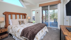 A bedroom with a skylight, a bed with a patterned bedspread, a wooden headboard, a nightstand with a lamp, and a sliding glass door leading to an outdoor area. - Ski-In/Ski-Out Chalet | Lone Peak Views | Private Hot Tub (Big Sky)