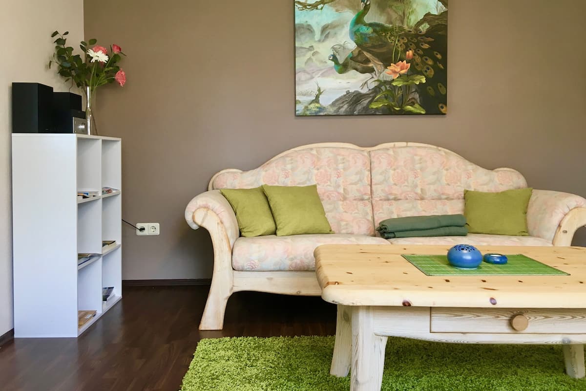 Un salon avec un canapé à motifs floraux, une table basse en bois, une bibliothèque et un tableau représentant un paon.