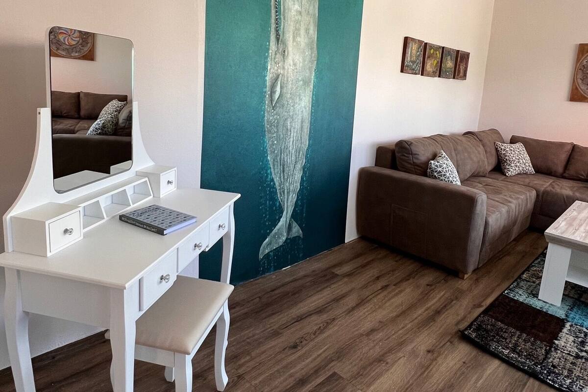 Un salon moderne avec un bureau blanc, un canapé marron et une grande fresque murale représentant une baleine.