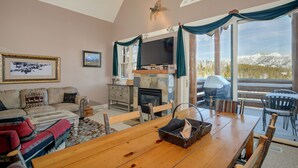 A cozy living room with a fireplace, a dining area with a wooden table, and a large window offering a mountain view. - Ski-in/ski-out home w/private hot tub in Moonlight Basin - Saddle Ridge - Montana Mountain Chalet (Big Sky)