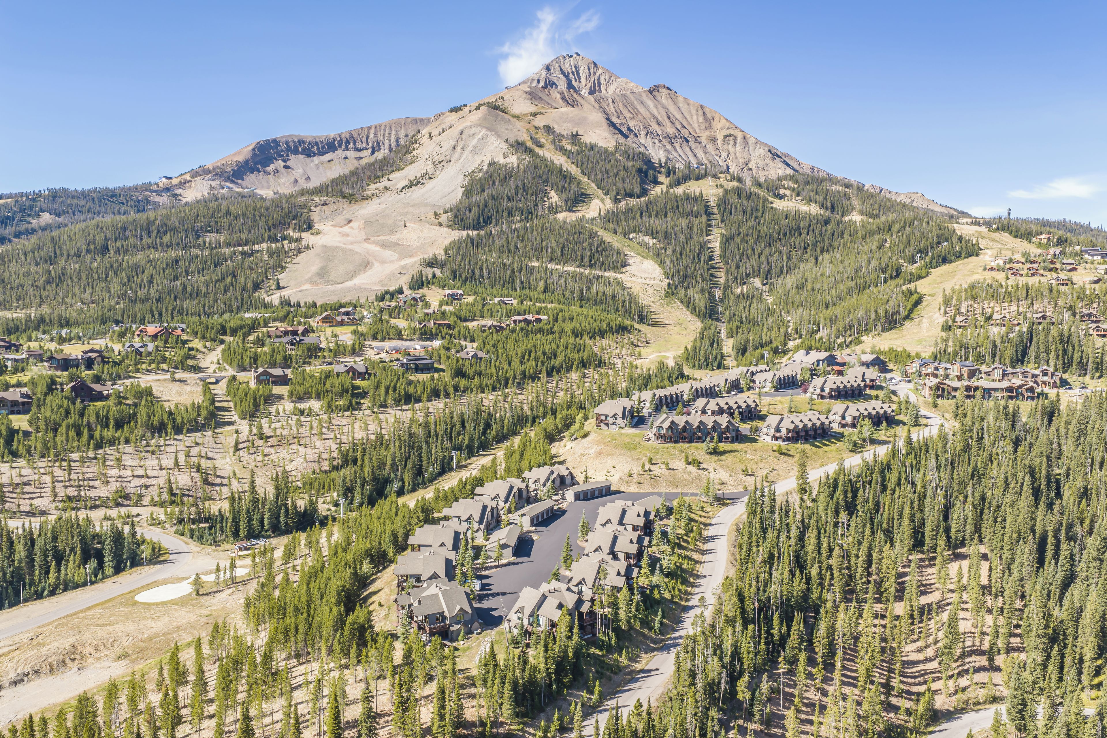 A mountainous landscape with a residential area featuring houses with sloped roofs, surrounded by dense forests and a clear sky.
