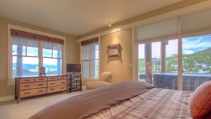 Une chambre à coucher avec un grand lit, une commode, un fauteuil et une vue sur un paysage montagneux, visible à travers de grandes fenêtres.