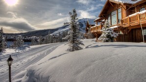 Una cabaña de madera con techo nevado, montañas al fondo y cielo despejado.