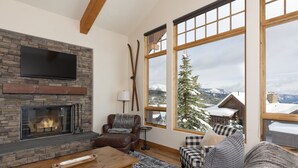 Un salón acogedor con chimenea de piedra, mobiliario de cuero y un amplio ventanal con vistas a una montaña nevada.