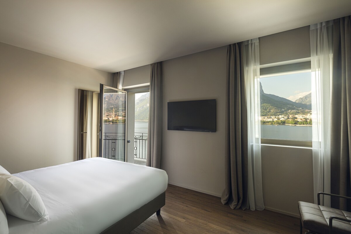 A hotel room with a bed, a TV mounted on the wall, and a view of a lake and mountains through large windows.