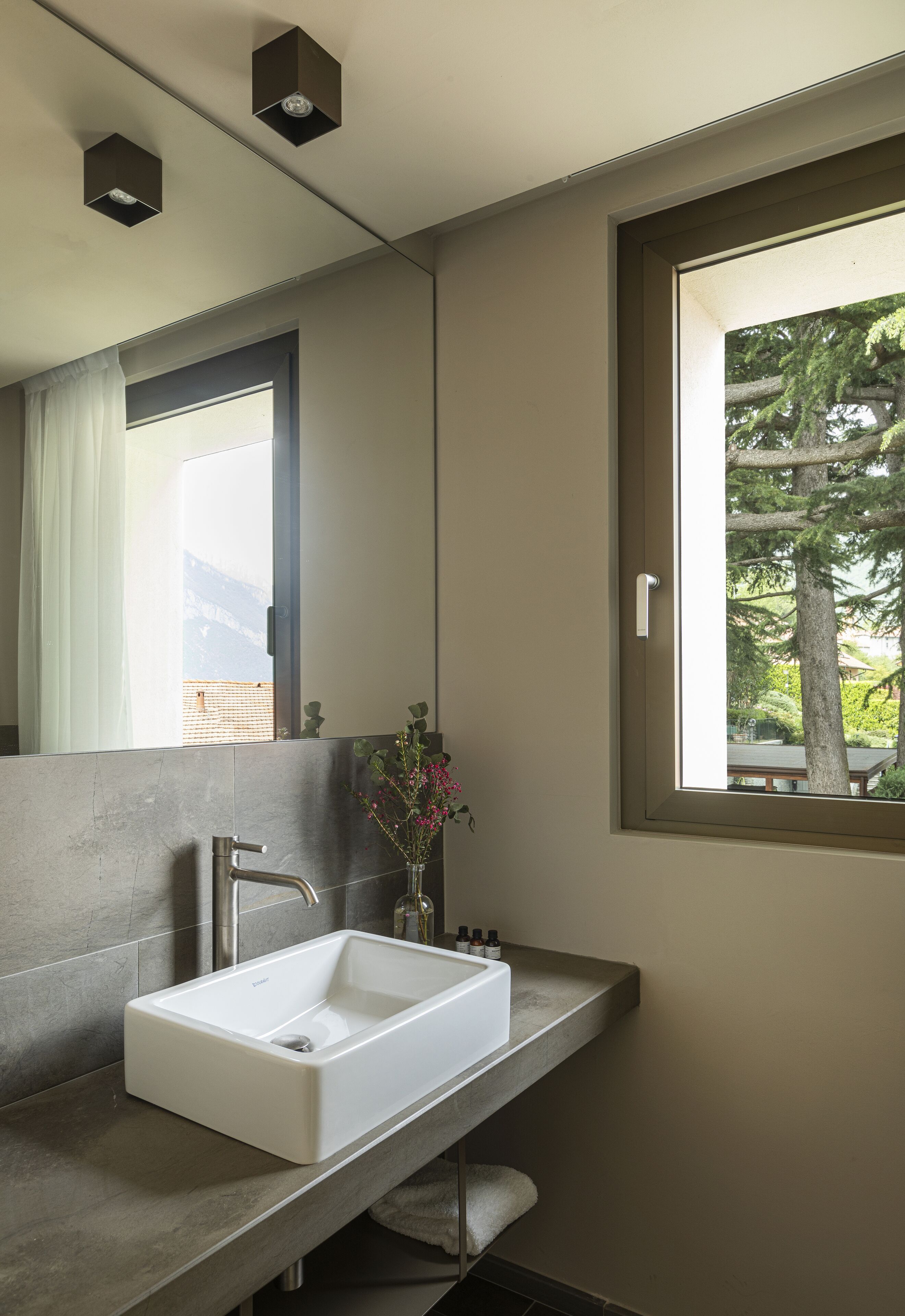A modern bathroom with a large mirror, a built-in sink, and a window offering a view of trees and a building.