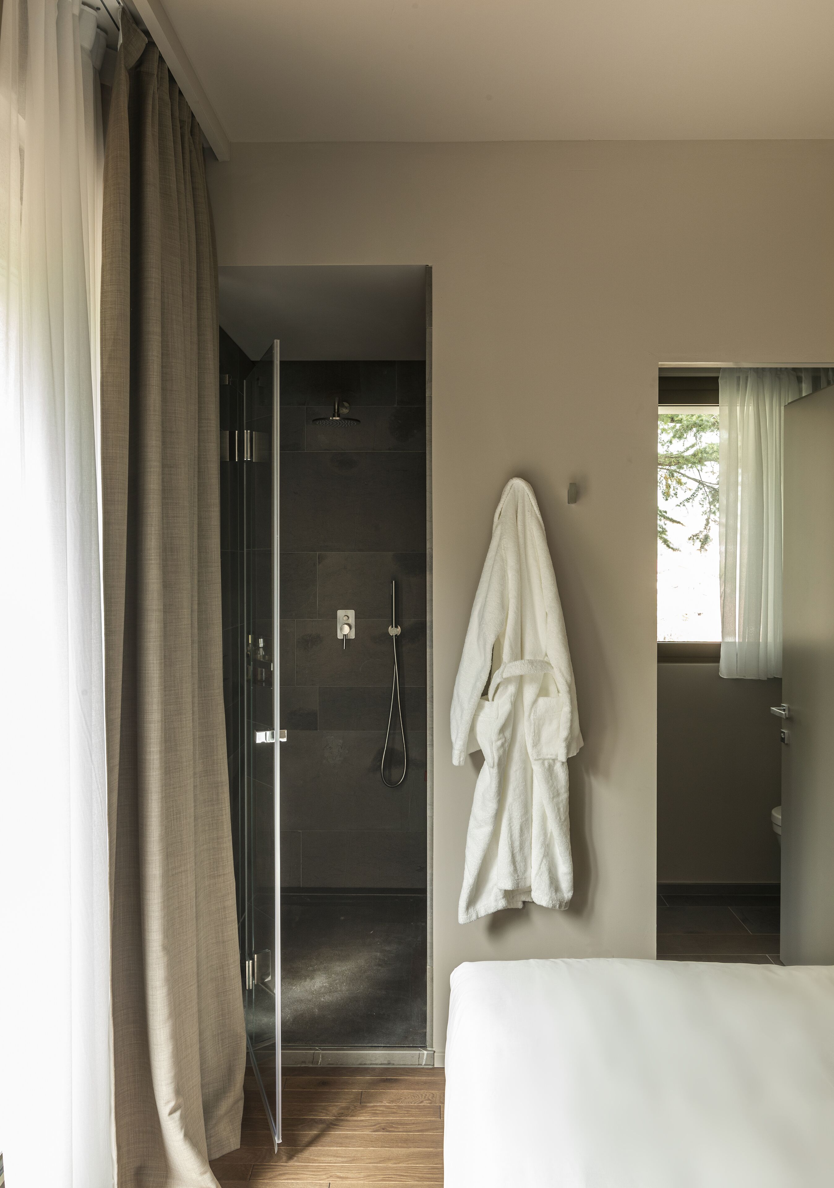 Une chambre d’hôtel moderne avec une porte de douche en verre, un peignoir blanc accroché au mur et un lit recouvert d’une couverture blanche.
