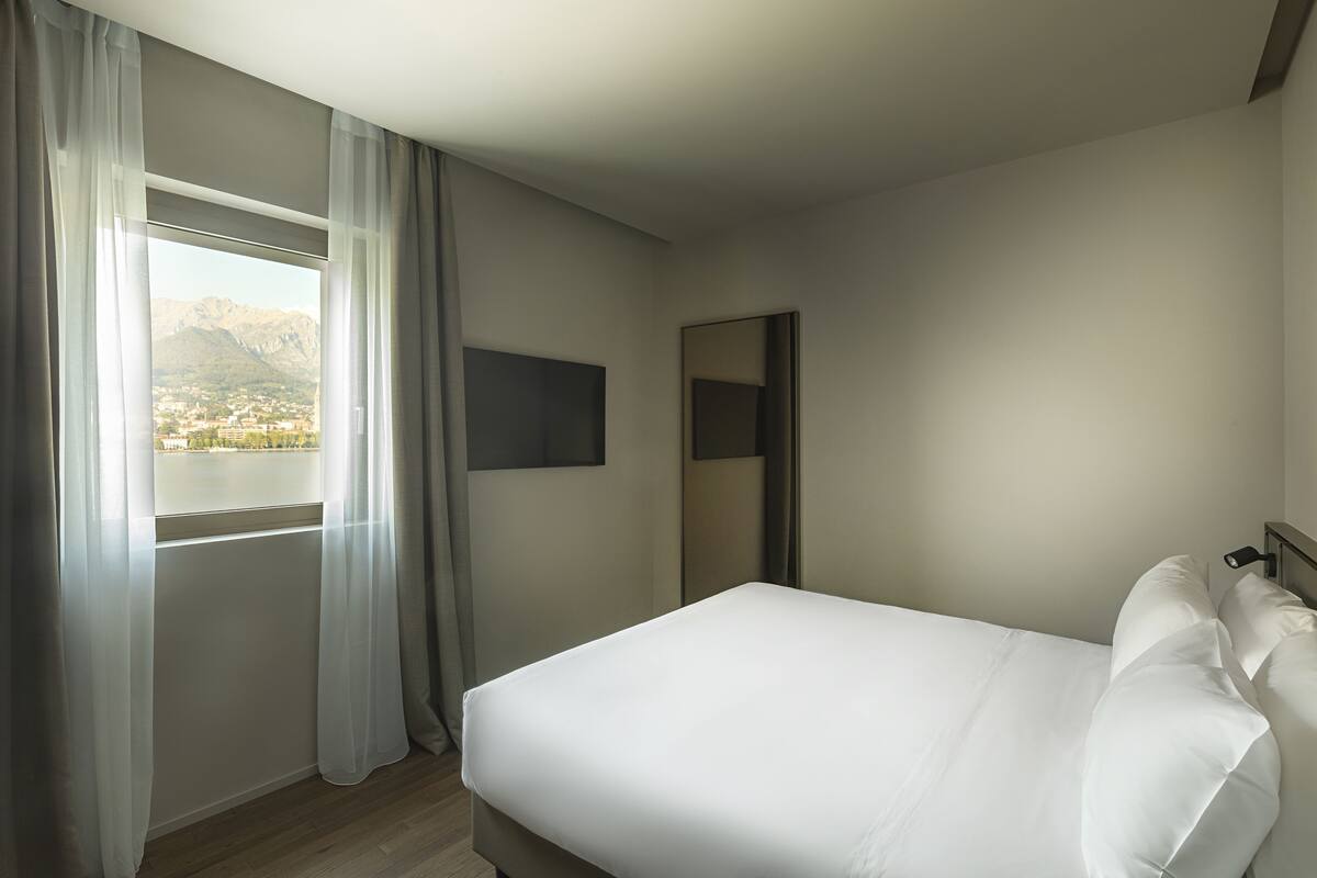 A hotel room with a bed, a window with curtains, and a flat-screen TV mounted on the wall.