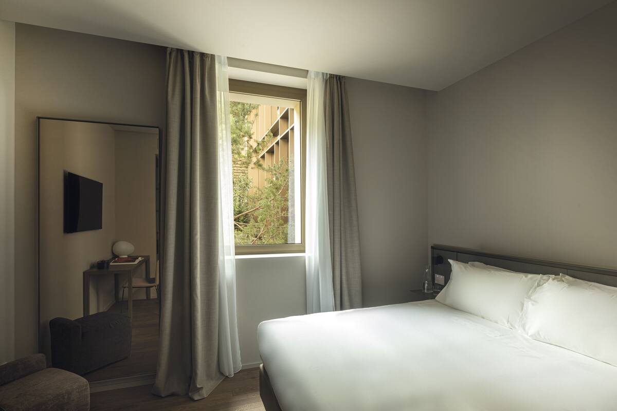 A bedroom with a large bed, a bedside table with a lamp, a chair, and a window with curtains.