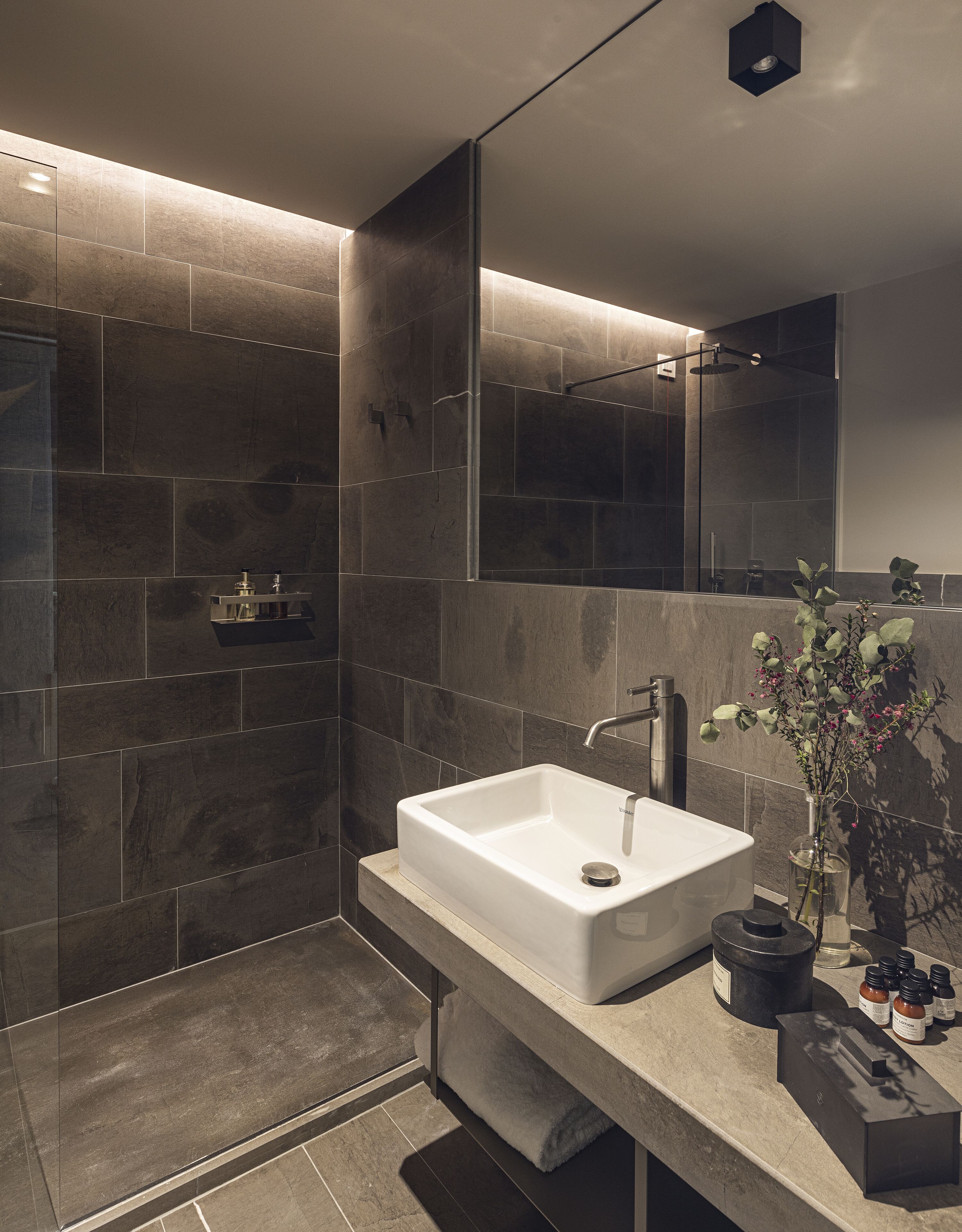 A modern bathroom with a large white sink, dark tiled walls, a glass shower enclosure, and a mirror reflecting the space.