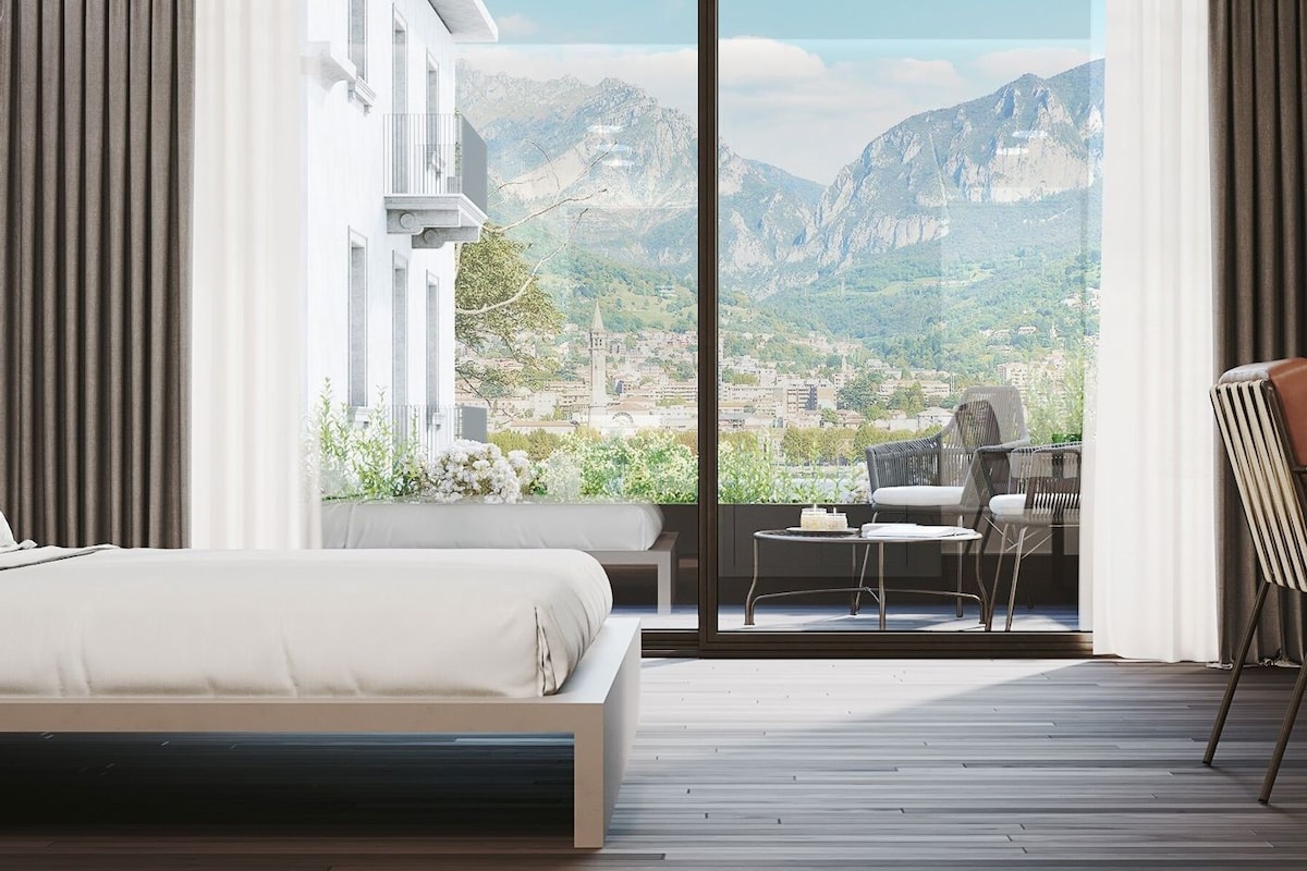 A modern hotel room with a bed, a desk, a chair, and a view of a mountainous landscape.