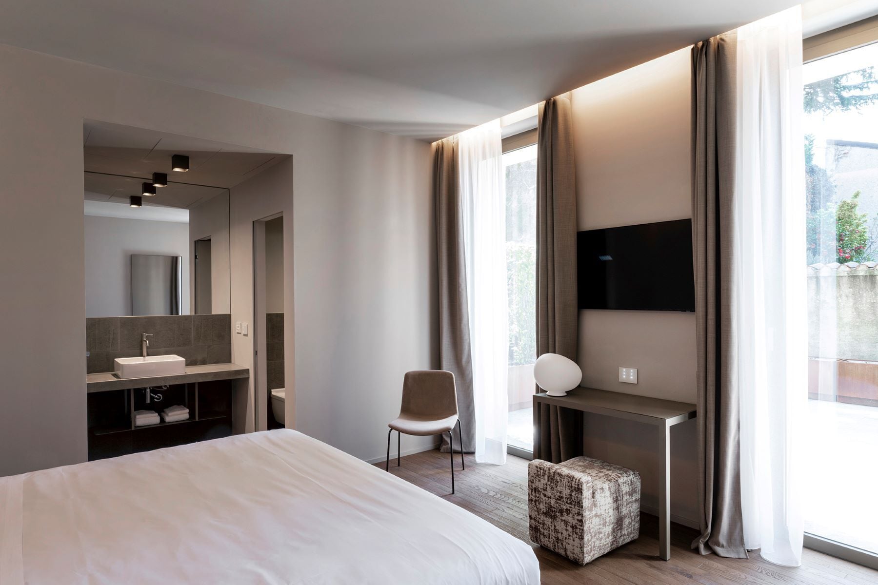 A modern hotel room with a large bed, a desk, a chair, a TV, and a bathroom with a mirror and sink.