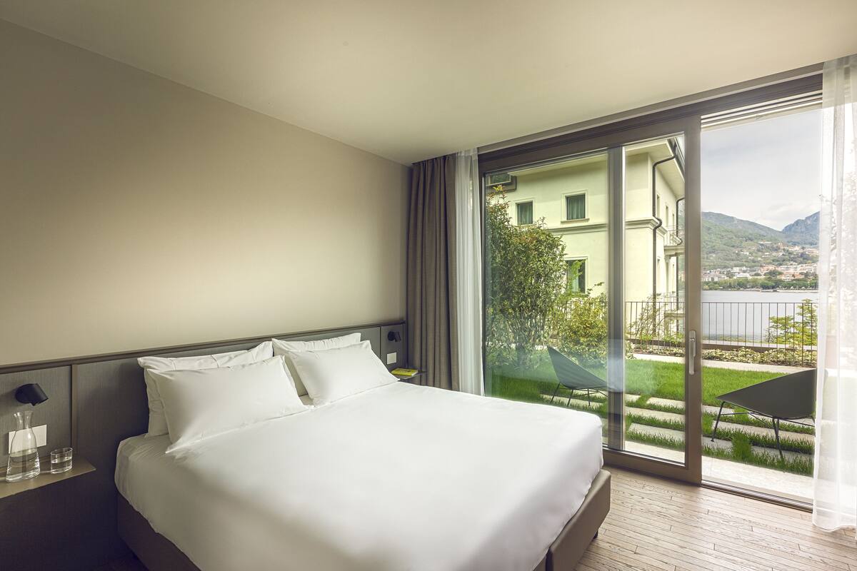 A hotel room with a large bed, a nightstand, a window with a view of greenery and buildings, and a sliding glass door to an outdoor area.