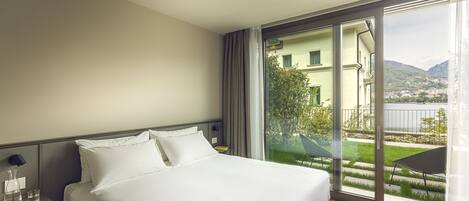 Habitación de hotel con una cama grande, una mesita de noche, un ventanal con vistas a zonas verdes y edificios, y una puerta corrediza de vidrio hacia un espacio al aire libre.