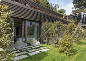 Una casa moderna con puerta de cristal, un pequeño patio con dos sillas y un jardín cuidado.