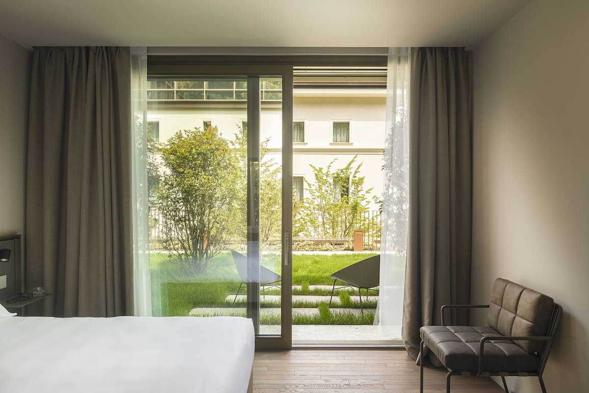 A hotel room with a bed, a chair, and a sliding glass door leading to a balcony with a view of greenery and a building.