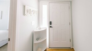 Un couloir étroit avec une porte blanche, une petite étagère blanche ornée d’objets décoratifs et un lit dans la pièce adjacente.