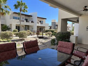 A patio area with a glass table and red patterned chairs, surrounded by a landscaped garden with various shrubs and palm trees, under a clear blue sky.