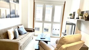 A living room with a sofa, armchair, and a coffee table. There is a window with a view of buildings outside.