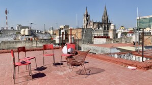 Una terraza en la azotea con sillas rojas, una maceta blanca y una silla de mimbre. Al fondo, se observa una catedral con dos torres y un edificio moderno con fachada de vidrio.