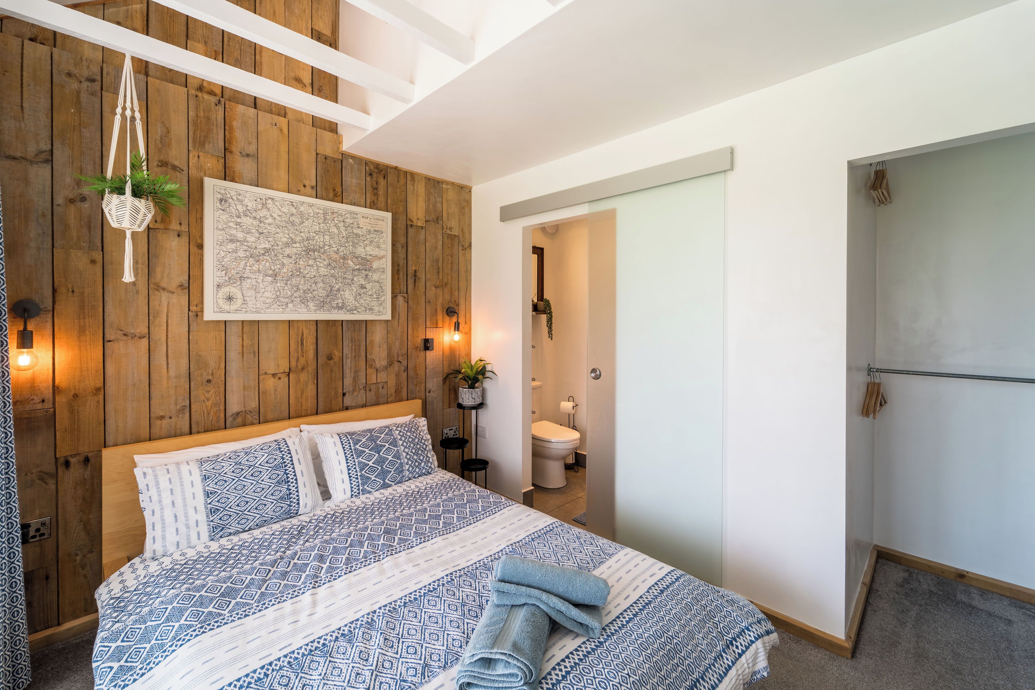 A bedroom with a wooden wall, a bed with patterned bedding, a hanging plant, and a map on the wall.