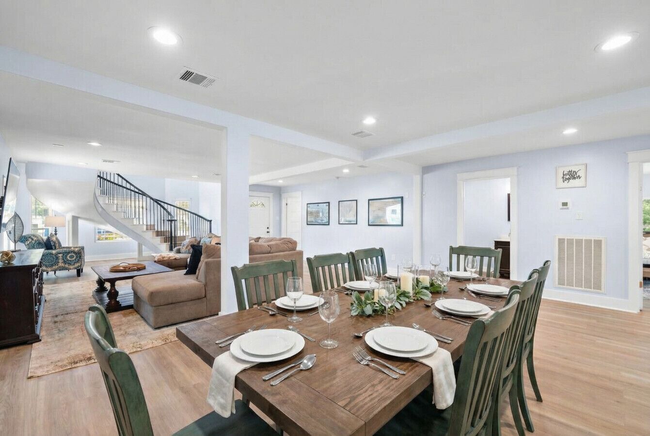 A spacious dining area with a wooden table set for six, surrounded by green chairs. A staircase is visible in the background leading to an upper level.